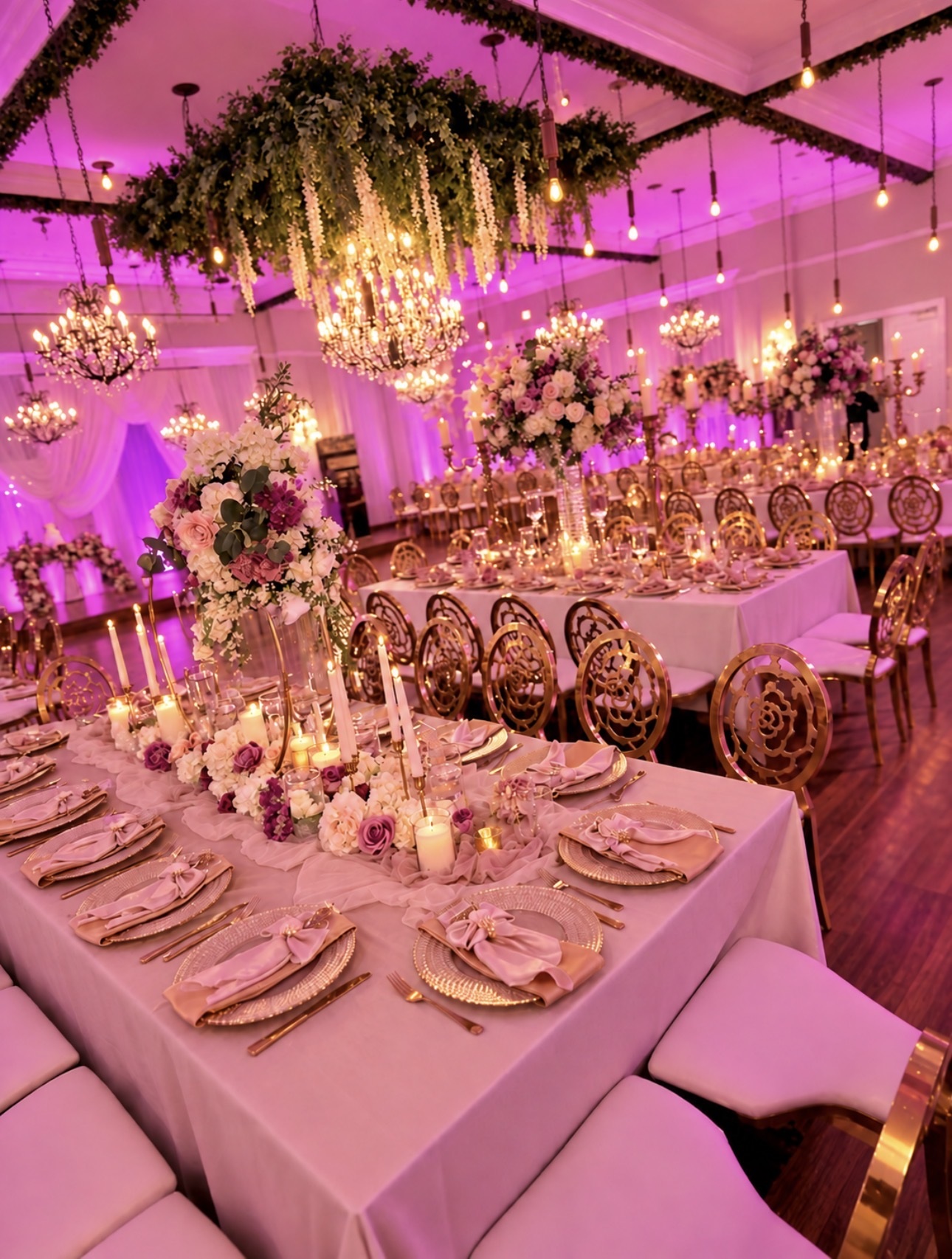 Pink-lit reception with chandeliers, floral centerpieces, gold chairs, and candlelit tables